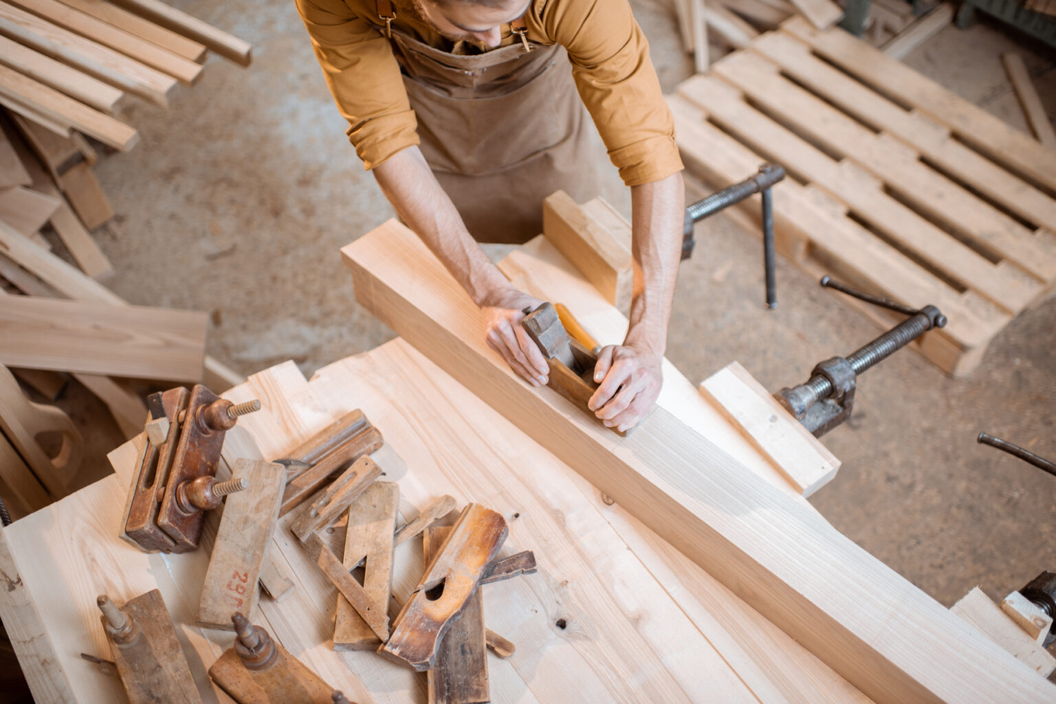 Handsome carpenter working with a wood, planing a bar with a plane on the worbench in the carpentry workshop. View from above